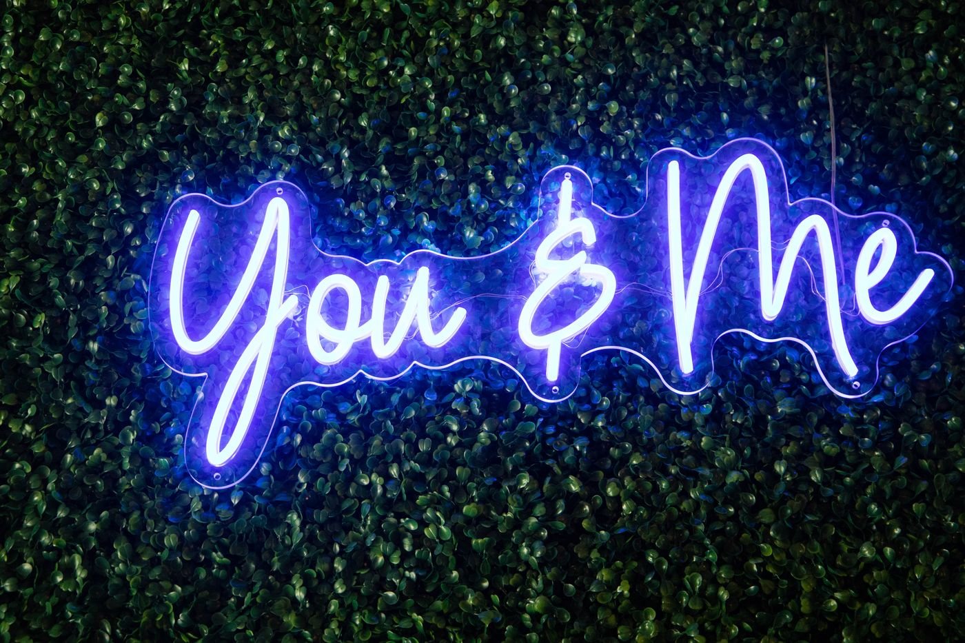 You and Me script neon sign glowing purple on a greenery wall